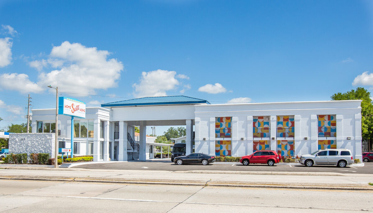 A white building with a blue roof and colorful windows, with a sign reading 'Home Suite Home' and several cars parked in front.